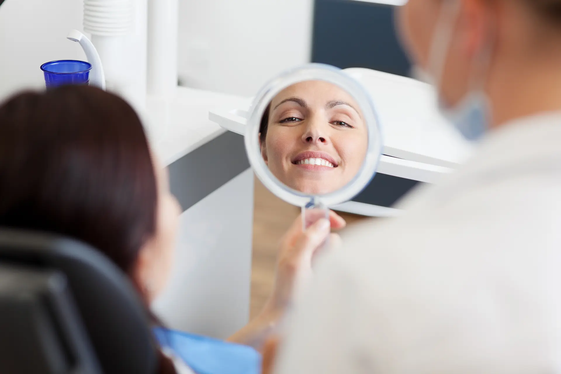 Patientin in Zahnarztpraxis begutachtet Zähne im Handspiegel, im Spiegel sieht man sie lächeln (Symbolbild für Ästhetische Zahnerhaltung im Frontbereich, Kronen und Verblendungen)